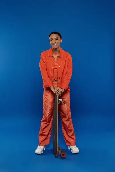 A young African American woman in an orange jumpsuit holding a skateboard on a vibrant blue background. — Stock Photo