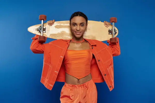 Vibrant young African American woman in orange outfit confidently holds skateboard overhead against blue backdrop. — Stock Photo