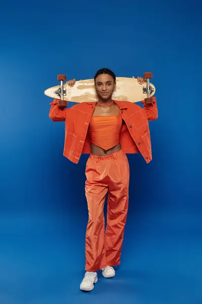 Young African American woman in vibrant orange outfit holds skateboard triumphantly over her head against a blue background. — Stock Photo