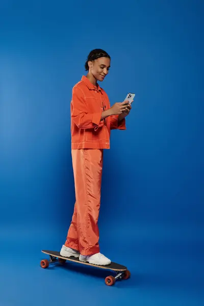 A vibrant young African American woman in an orange outfit stands on a skateboard, looking at her cell phone. — Stock Photo