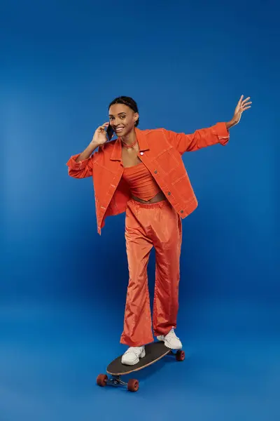 A young African American woman in a bright orange suit gracefully skateboarding against a vibrant blue background. — Stock Photo