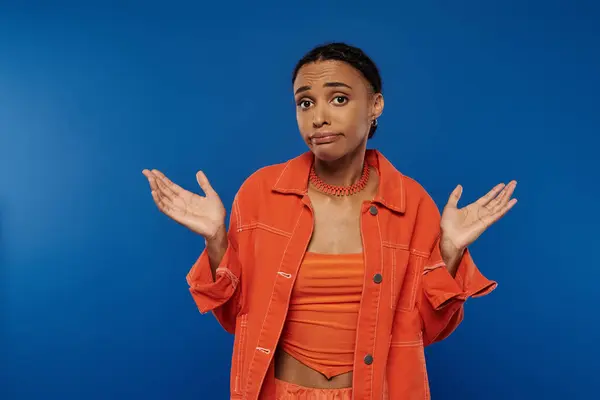 Confused African American woman in an orange shirt gesturing against a bright blue background. — Stock Photo