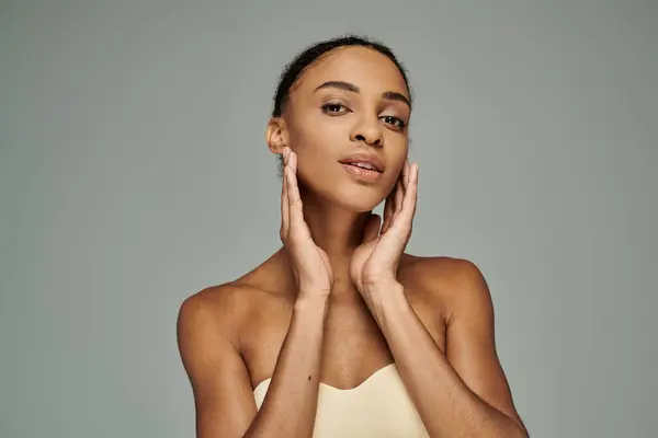 A beautiful young African American woman in a strapless top, gently touching her face with both hands, on a grey background. — Stock Photo