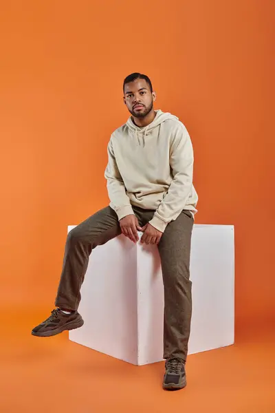 Stylish African American man sits atop white block amidst vibrant backdrop. — Stock Photo