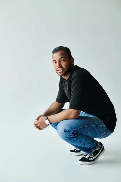 Um homem afro-americano de camisa preta e jeans ajoelha-se em um cenário vibrante. — Fotografia de Stock