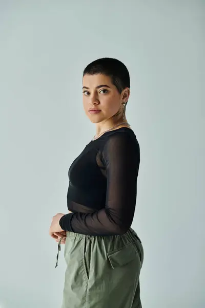 Young woman with short hair posing confidently in stylish black top and green pants against a grey background. — Stock Photo