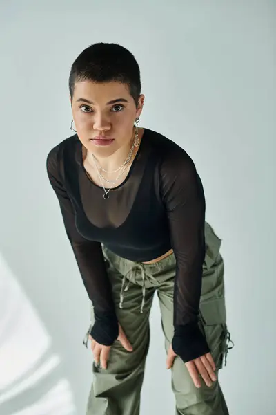 Young woman with short hair strikes a pose in fashionable black shirt and green pants on a grey background. — Stock Photo