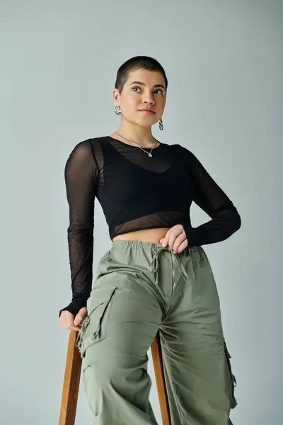 Kurzhaarige junge Frau in stylischer Freizeitkleidung sitzt auf einem Stuhl, die Hände auf den Hüften und strahlt Zuversicht und Durchsetzungsvermögen aus. — Stockfoto