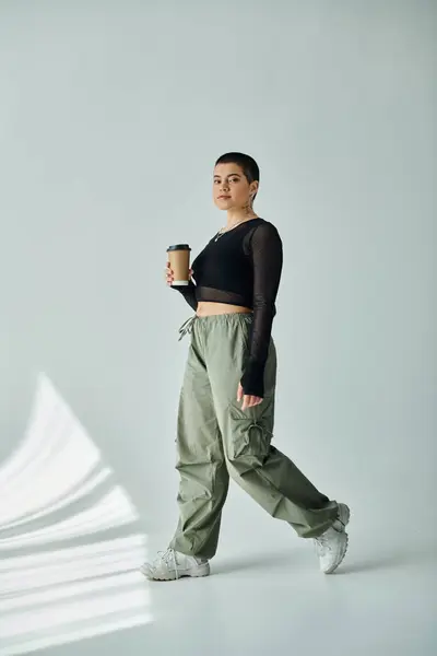 Una joven elegante con el pelo corto camina con gracia, taza de café en la mano, sobre un fondo gris. - foto de stock