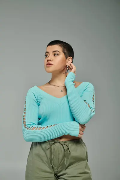 Eine stylische junge Frau mit kurzen Haaren posiert in blauem Pullover und grüner Hose vor grauem Hintergrund. — Stockfoto