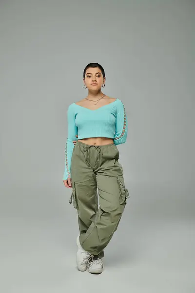 Una mujer joven y elegante con el pelo corto posando con confianza en un top de moda y pantalones de carga sobre un fondo gris. — Stock Photo