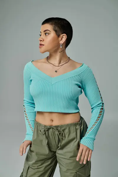 A young woman with short hair poses confidently in a green sweater and cargo pants against a grey background. — Stock Photo