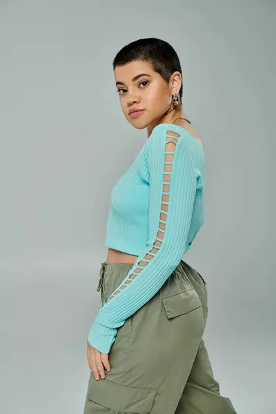 Young woman with short hair strikes a pose in a stylish blue sweater and khaki pants against a grey backdrop. — Stock Photo