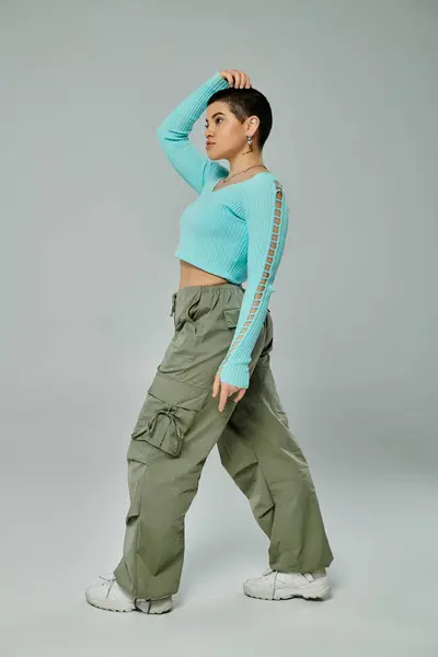 Young woman with short hair striking a pose in a stylish outfit of a blue top and green pants on a neutral grey background. — Stock Photo