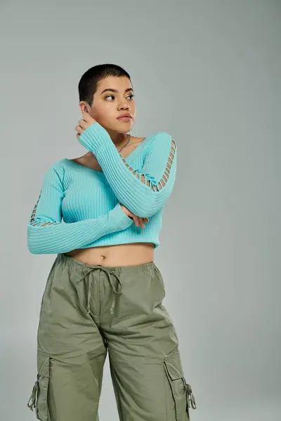 A young woman with short hair poses stylishly in green pants and a blue sweater against a grey background. — Stock Photo