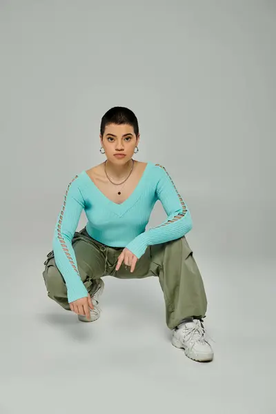 A stylish young woman with short hair posing in a blue shirt and green pants on a grey background. — Stock Photo