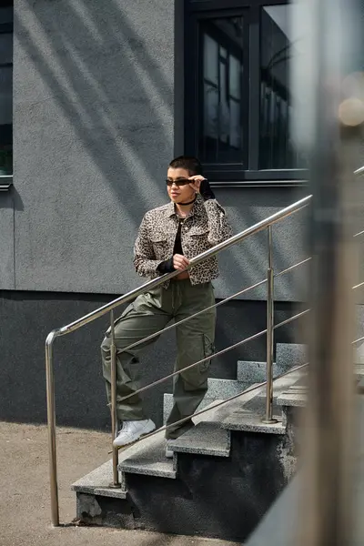 Молодая женщина в стильной одежде стоит на лестнице рядом с современным зданием. — стоковое фото