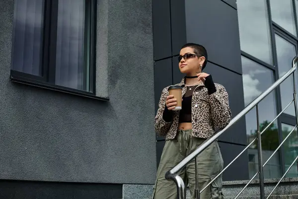 A stylish young woman with short hair and a leopard print jacket, standing on a staircase, enjoying a cup of coffee. — Stock Photo