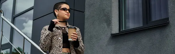 A young woman with short hair wearing a leopard print jacket, holding a drink in front of a modern building. — Stock Photo