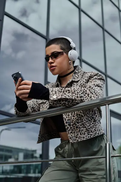 A young woman with short hair and a leopard print jacket listens to music on headphones while holding a cell phone. — Stock Photo
