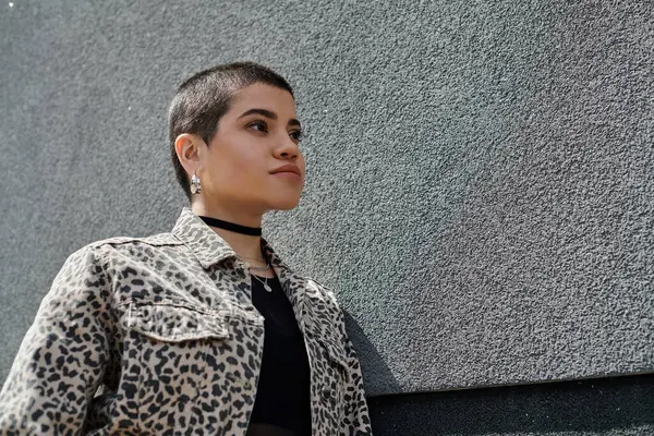 A stylish young woman with short hair stands against a concrete wall, showcasing a trendy leopard print jacket. — Stock Photo