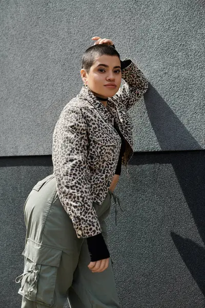 A stylish young woman in a leopard print jacket leans against a modern building, hand on head. — Stock Photo
