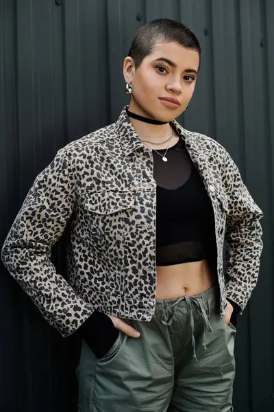Una mujer joven y elegante con el pelo corto se para con confianza delante de una elegante pared negra, exudando elegancia urbana. - foto de stock