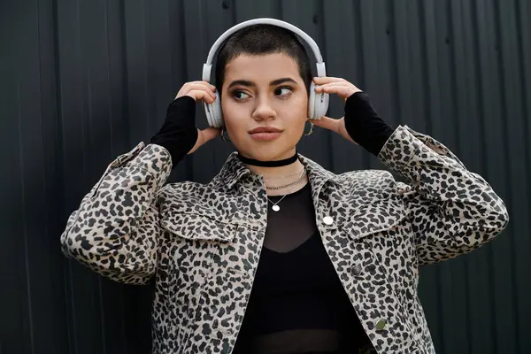 A young woman with short hair wearing headphones and a leopard print jacket stands near a modern building. — Stock Photo