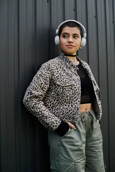 Une femme élégamment habillée aux cheveux courts se tient à côté d'un mur moderne, portant des écouteurs. — Photo de stock