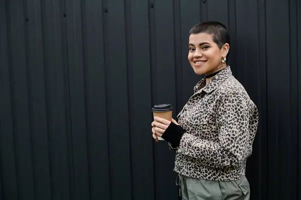 A young woman in chic attire holding a cup of coffee, standing in front of a sleek black wall. — Stock Photo