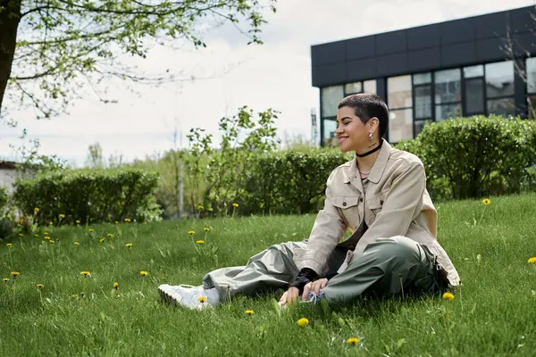 Young woman with short hair sits on grass in front of a building, lost in thought. — Stock Photo