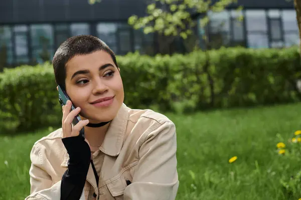 A young woman with short hair smiling as she talks on smartphone while sitting on green grass outdoors. — Stock Photo