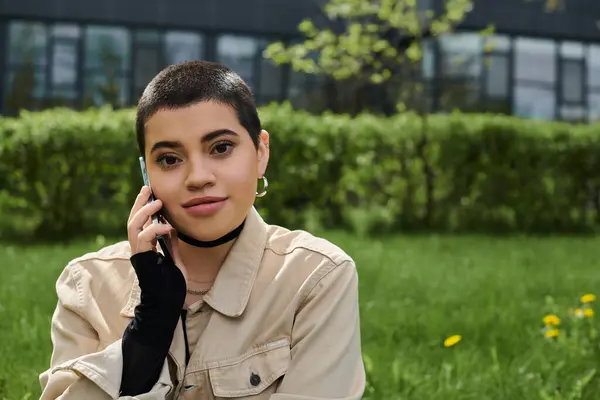 A young woman with short hair in stylish casual attire sits in the grass, talking on her cell phone. — Stock Photo