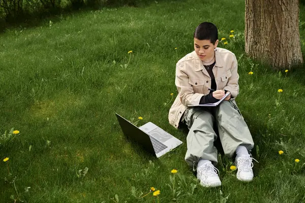 A young woman with short hair, in stylish casual attire, sitting on green grass outdoors, working on a laptop. — Stock Photo