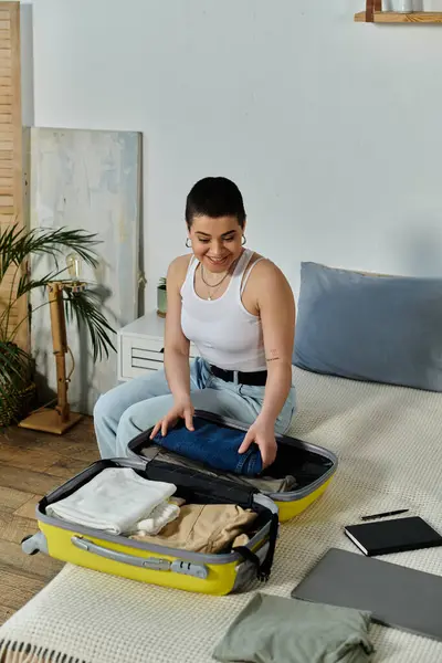 A young woman with short hair meticulously folds clothes into her open suitcase on a cozy bed, preparing for an upcoming trip. — Stock Photo