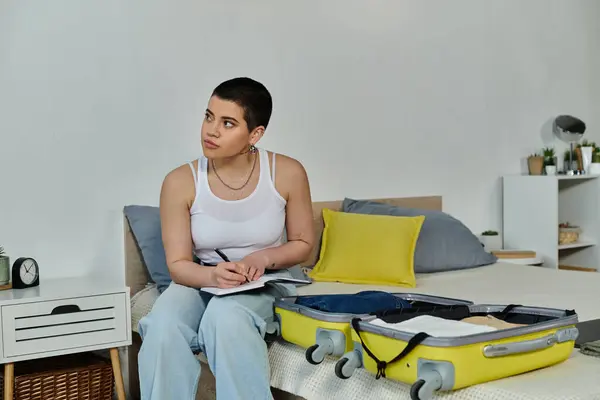 A young woman with short hair sitting on a bed, packing a yellow suitcase for an upcoming vacation and trip. — Stock Photo