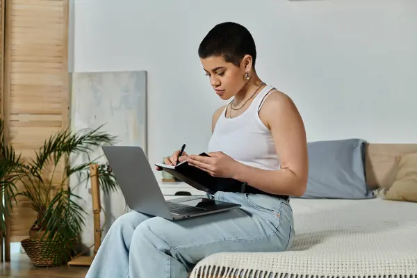 A young woman with short hair in casual wear sits on a bed, engrossed in using a laptop computer. — Stock Photo