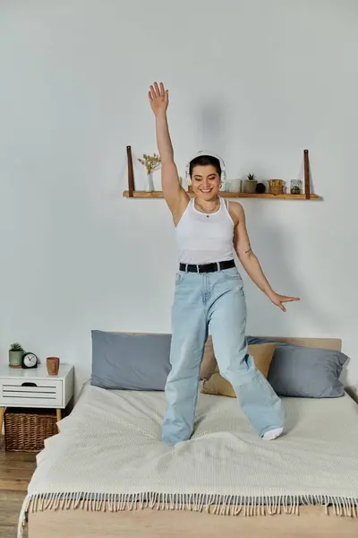 A young woman with short hair and casual clothes standing on a bed with arms raised in the air, expressing joy. — Stock Photo
