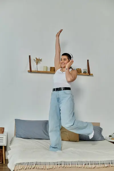 A young woman with short hair joyfully stands on a bed with arms raised in the air. — Stock Photo