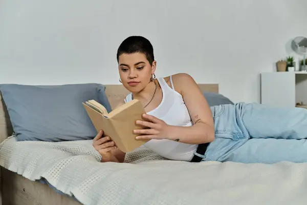 A young woman with short hair, dressed in casual wear, lies peacefully on a bed, immersed in reading a book. — Stock Photo