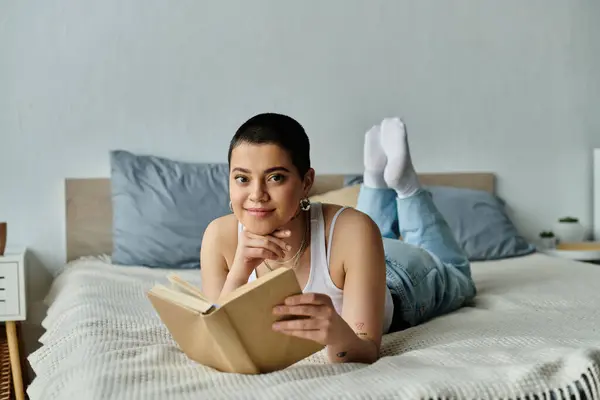 A young woman with short hair, dressed casually, lays on a bed engrossed in reading a book. — Stock Photo