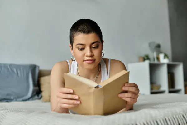 A young woman with short hair in casual wear reclining on a bed, engrossed in reading a book. — Stock Photo