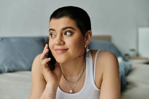 A young woman with short hair in casual wear sitting on a bed, talking on a cell phone. — Stock Photo
