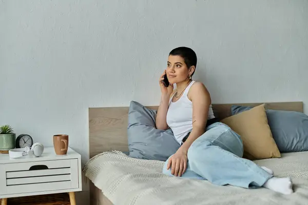 A young woman with short hair in casual wear sitting on a bed, engaged in a phone call. — Stock Photo