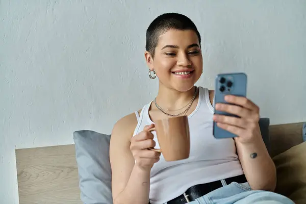 A young woman with short hair sits on a bed, engrossed in her cell phone, wearing casual attire. — Stock Photo