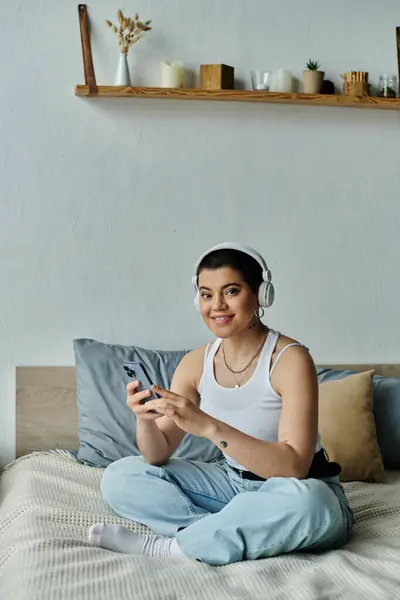 A young woman with short hair sits on a bed, absorbed in her phone screen. — Stock Photo