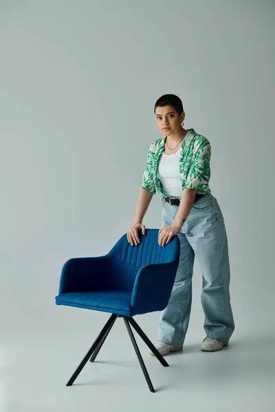 Una giovane donna elegante con i capelli corti si erge con grazia accanto a una sedia blu moderna in un ambiente domestico accogliente. — Foto stock