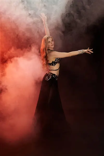 A mesmerizing young woman in a black dress gracefully dances amidst swirling smoke. — Stock Photo
