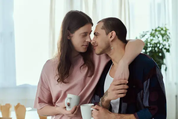 Una pareja gay se abraza calurosamente, cada uno sosteniendo una taza de café, mirando amorosamente el uno al otro en un ambiente acogedor en casa. - foto de stock