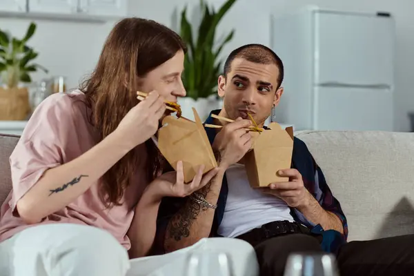 A casual gay couple enjoying a meal together on a couch at home. — Stock Photo